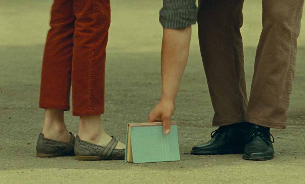 A closeup on two pairs of feet, and one of their owners bending down to pick up a book from the floor, in What Do We See When We Look at the Sky?, one of the best films of Berlinale 2021.