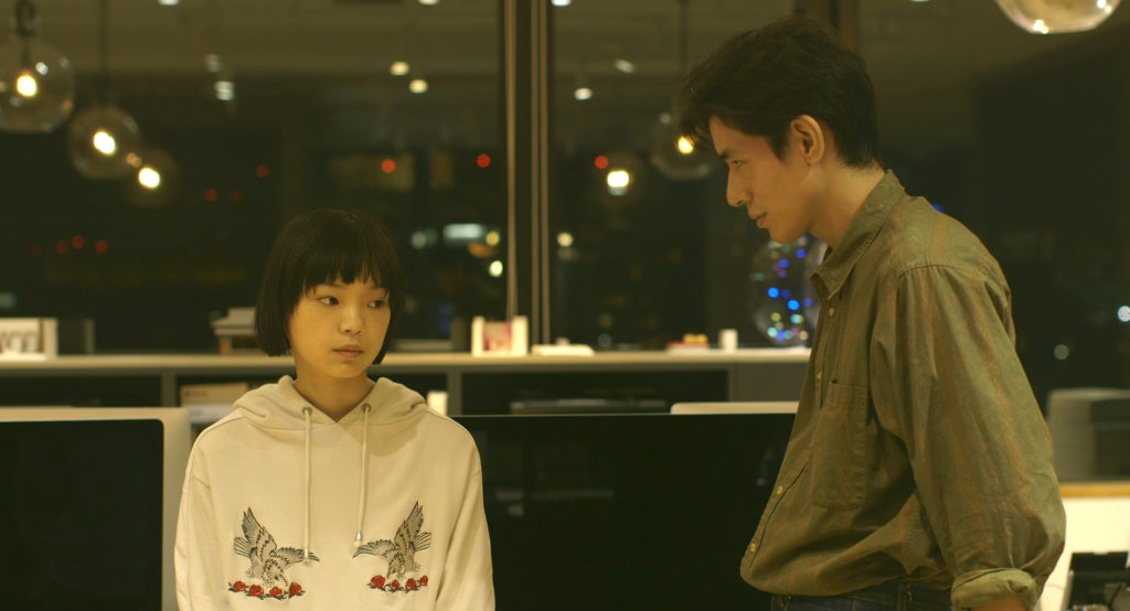 A young woman and a tall man stand next to each other, not quite like at each other, in Ryûsuke Hamaguchi's Wheel of Fortune and Fantasy.