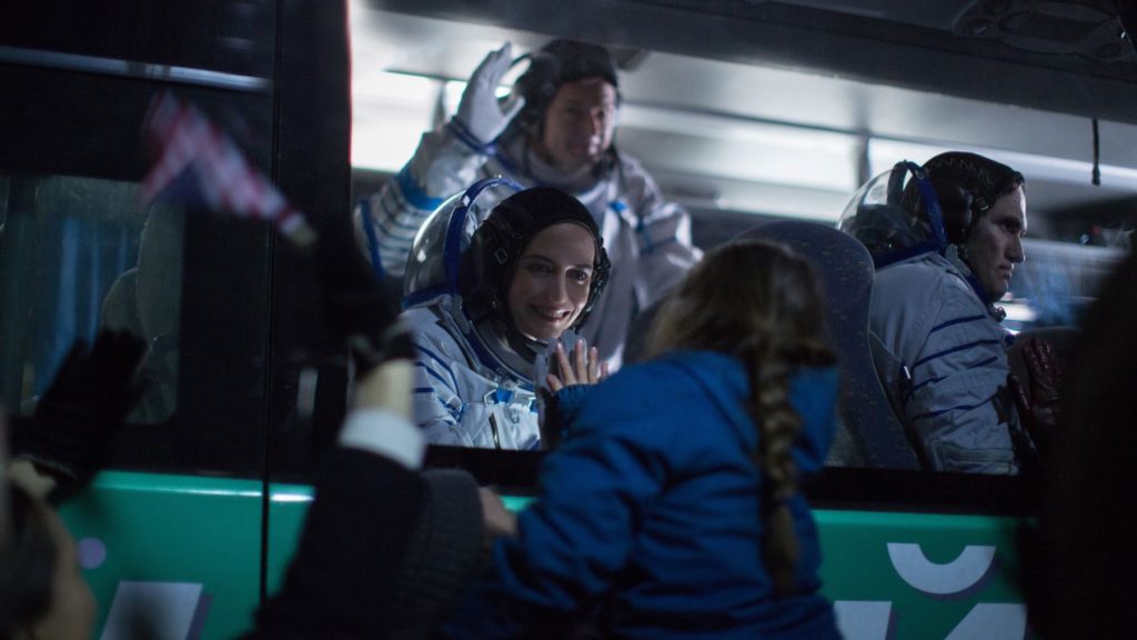 Eva Green stars as Sarah in Proxima (2019) directed by Alice Winocour. Sarah, dressed in her flight suit, is saying goodbye to her daughter through the window of the bus. They are touching hands against the glass.