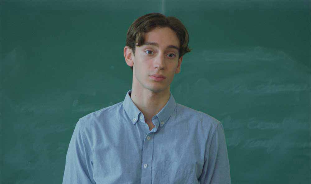 A mid shot of Théodore Pellerin in Genèse, standing against a green school board, dressed in blue school uniform. Pellerin also stars in Continental Drift (South), a film by Lionel Baier.