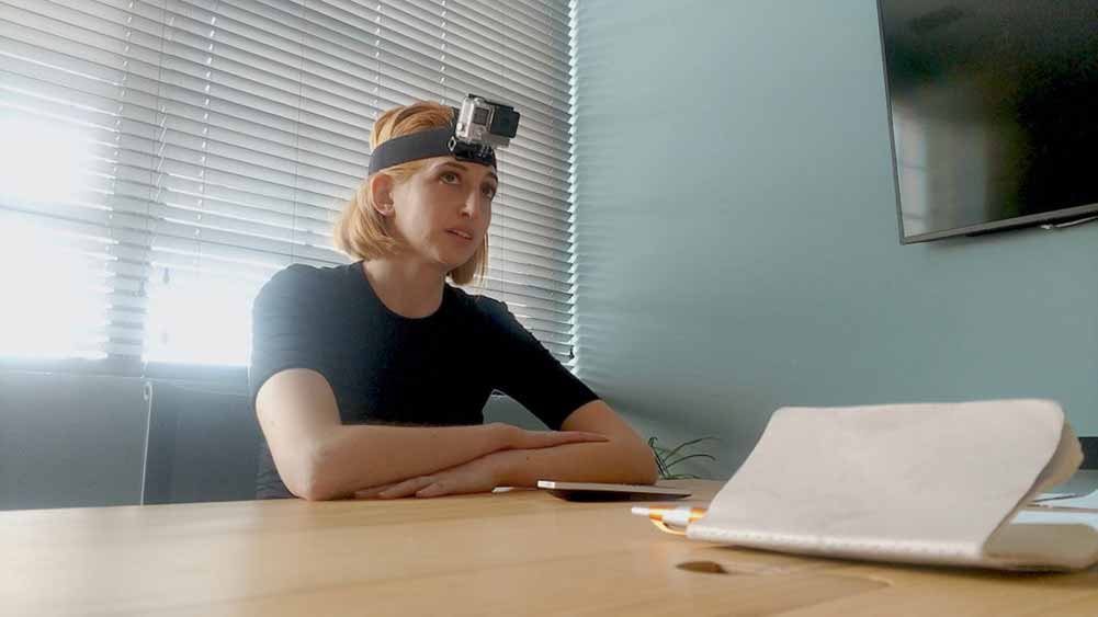 A woman with a camera strapped to her head sits at a board table in I Blame Society.