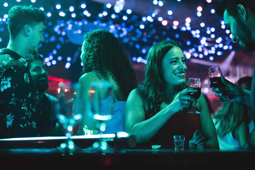 A still from Nadia, Butterfly in which Nadia clinks drinks with a man in a club, smiling.