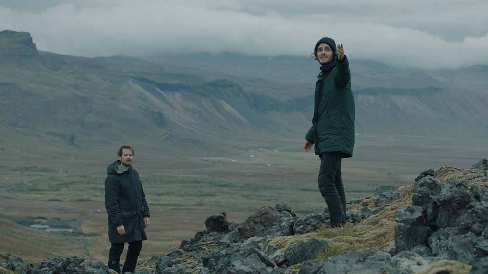 A still from Everything in the End, in which two people stand near to each other against a majestic Icelandic landscape.