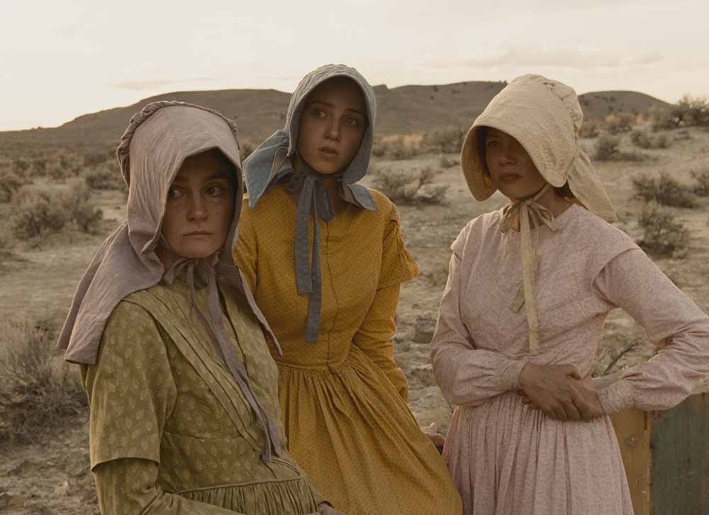 Three women in bonnets stand against a desert landscape in Meek's Cutoff.