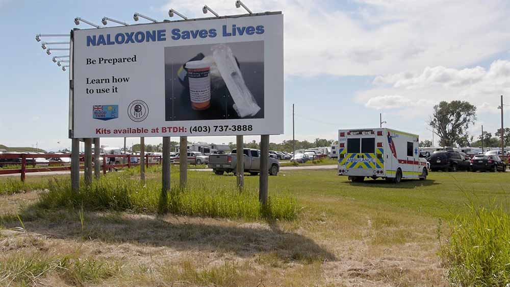 A still of a billboard that reads 'Naloxone saves lives' and features a picture of the drug.