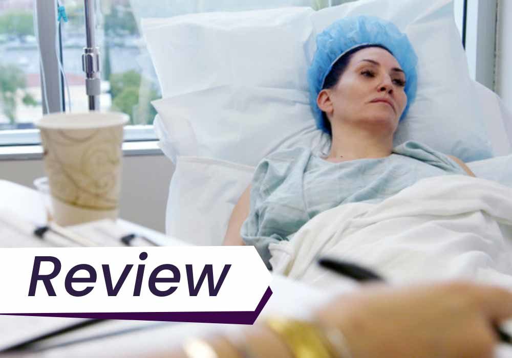A still from the film Explant, which shows Michelle Visage lying in a hospital bed, recovering from surgery. The text in front of the image reads, "Review".