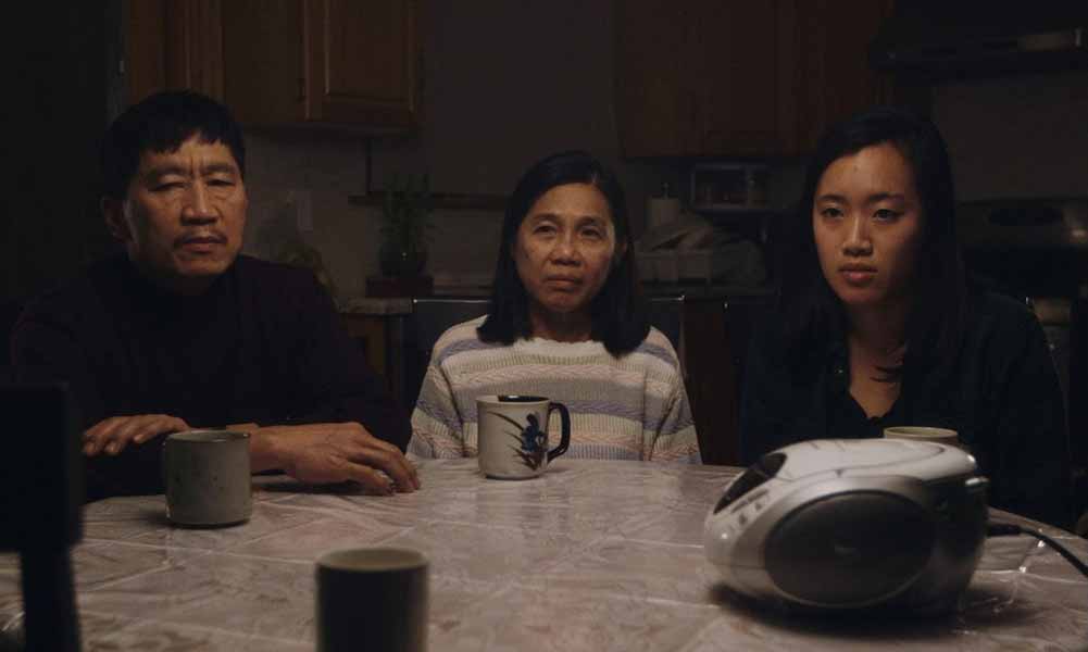 A still from No Crying at the Dinner Table, in which a father, mother, and daughter sit somberly around a kitchen table, listening to a tape recorder.
