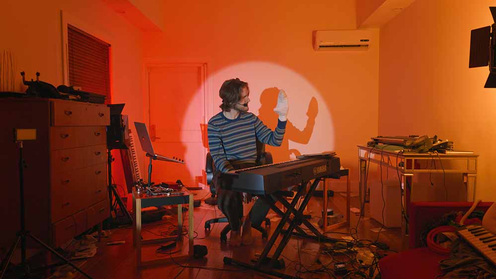 A still from Bo Burnham: Inside in which he sits at his piano in front of an orange-lit backdrop, holding a sock puppet on his hand.