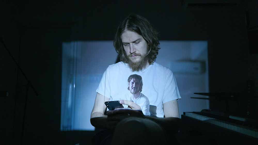A still from Bo Burnham: Inside in which a video of Burnham is projected onto his white t-shirt, as he watches it back on his phone.