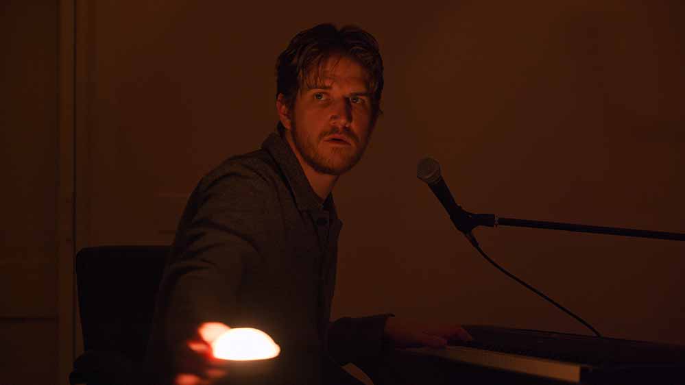 A still from Bo Burnham: Inside in which Burnham sits at a piano, holding a small light in front of his face to test out how it looks on camera.