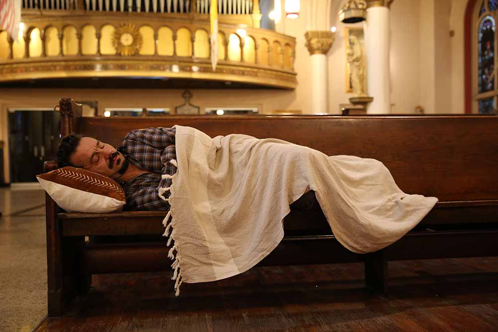 A still in which Paul lies down on the church pews, under a thin white blanket, his head resting on a small white pillow.