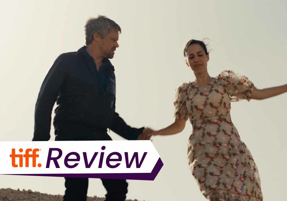 A still from Ahed's Knee in which Y, a middle aged man, holds Yahalom's hand, a young woman in a dress, as they walk up a hill. The text on the images reads, 'TIFF Review'.