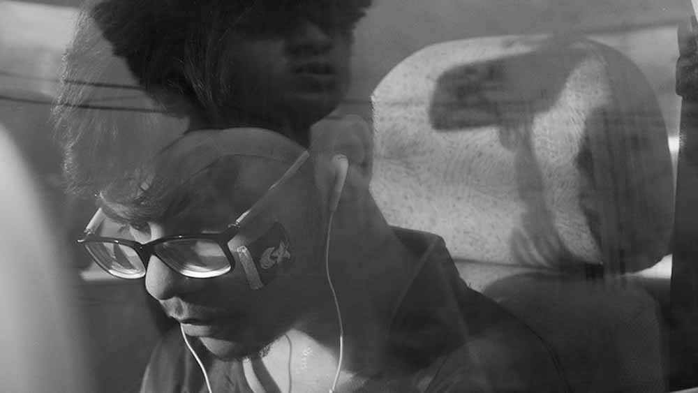 A black and white still of a young man in a car, seen through the car window, and another man reflected in the window.