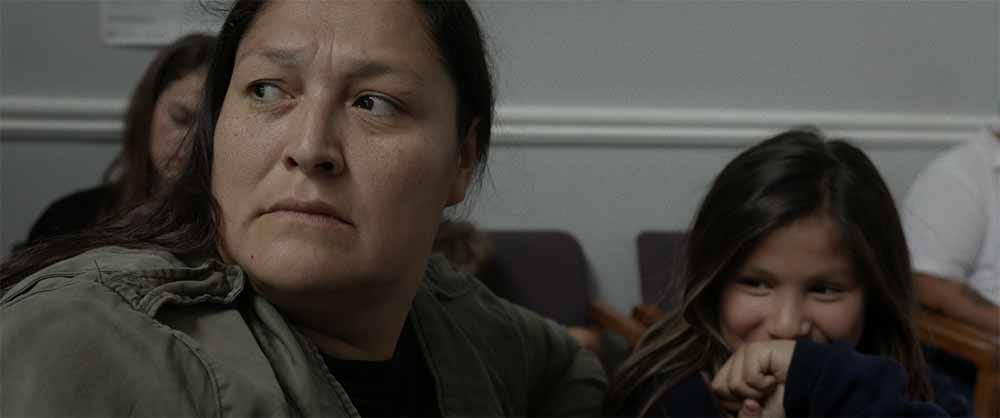 A still of a worried Indigenous mother with her young daughter, who is giggling, in a doctor's waiting room.