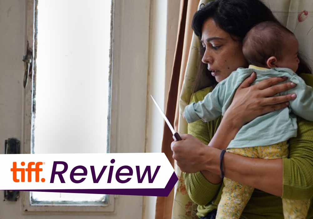 A still from Huda's Salon, in which a woman clasps her baby to her chest and holds out a knife, hiding in anticipation of attack. The text on the image reads, 'TIFF Review'.