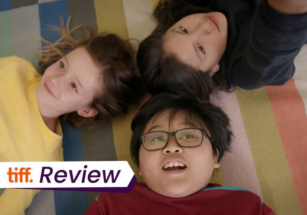 A still from Scarborough in which three young kids lie down on a colourful floor, gazing happily at the ceiling. The text on the image reads 'TIFF Review'.
