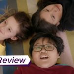 A still from Scarborough in which three young kids lie down on a colourful floor, gazing happily at the ceiling. The text on the image reads 'TIFF Review'.