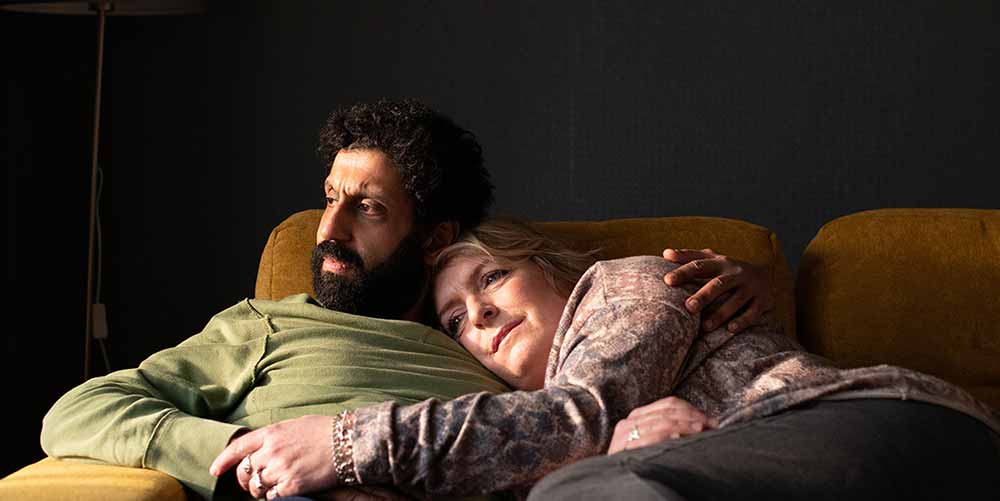 A still from Ali & Ava, in which the couple lies on a yellow sofa, Ava leaning on Ali's chest as he looks out the window in worry.