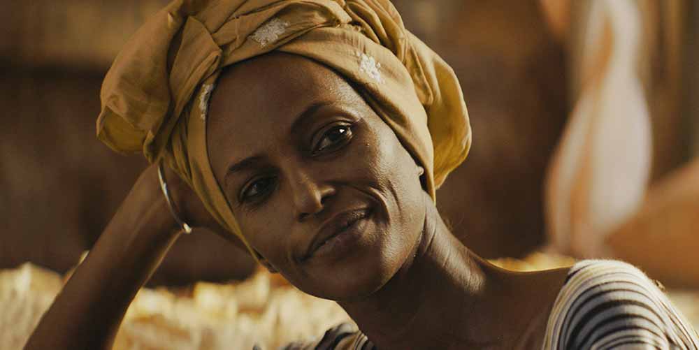 A still from Khadar Ayderus Ahmed's The Gravedigger's Wife, a closeup of the character Nasra, a Black woman wearing a yellow headwrap, looking contentedly off screen.