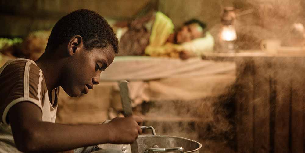 A still from Khadar Ayderus Ahmed's The Gravedigger's Wife, in which a young boy, Mahad, stirs a pot of food in the foreground, while his mother lies down on a bed, out of focus, in the background.