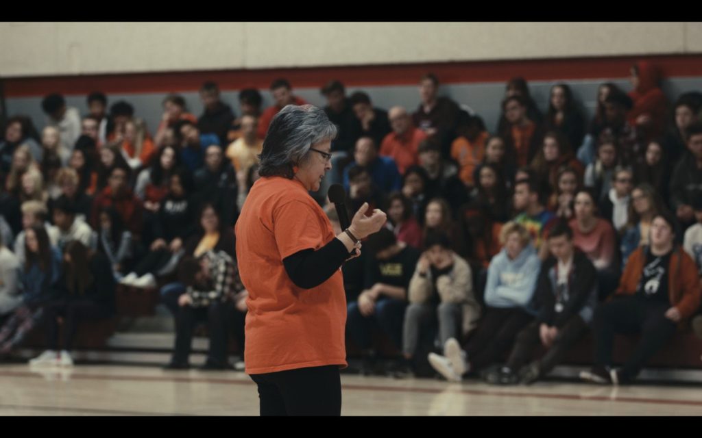 Still of Phyllis Jack-Webstad in an orange t-shirt talking at a high school about residential schools for Orange Shirt Day in Returning Home