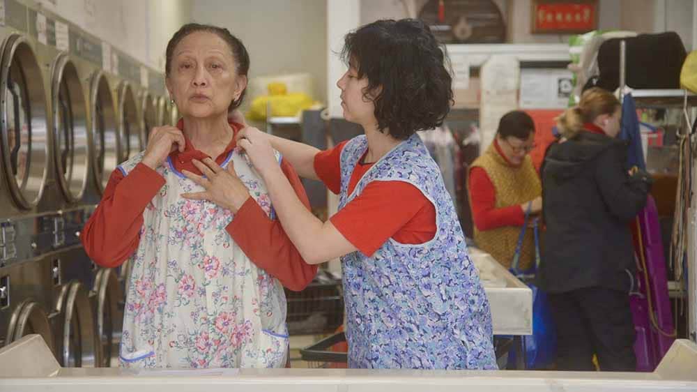 Two women, one older, the other younger, stand in the laundromat where they work, the younger woman fixing the older woman's apron.