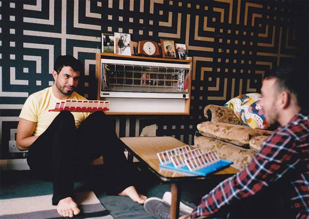 A still from Weekend, in which Russell and Glen sit on Russell's floor playing Guess Who, with a modern, patterned wallpaper behind them. The production design is by Sarah Finlay.