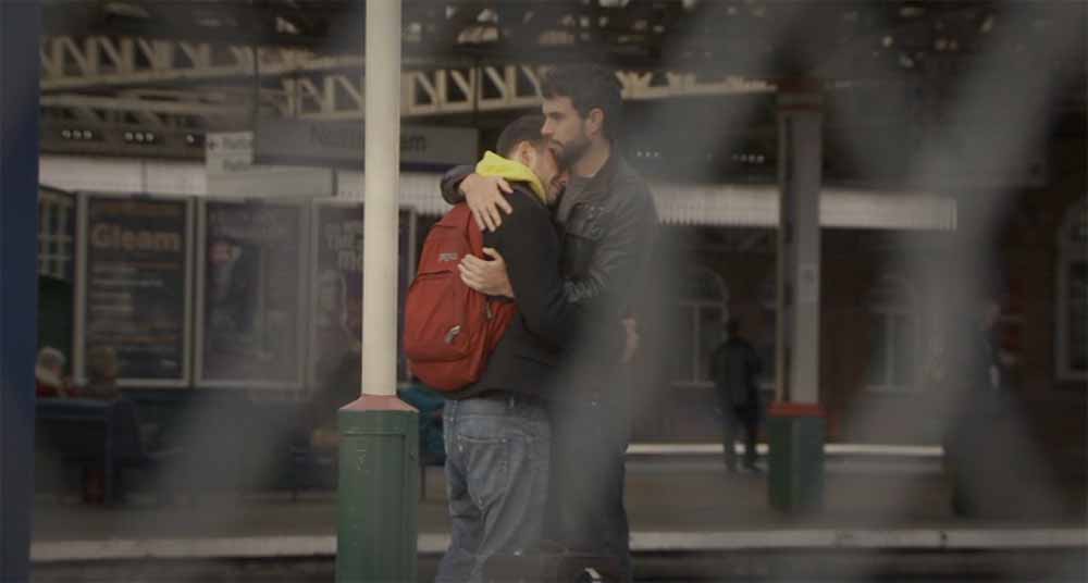 A still from Weekend, in which Russell and Glen, two young white men, embrace at a train station. In the foreground of the image, which was shot by Ula Pontikos, is a mesh gate in soft focus which the scene is shot through.