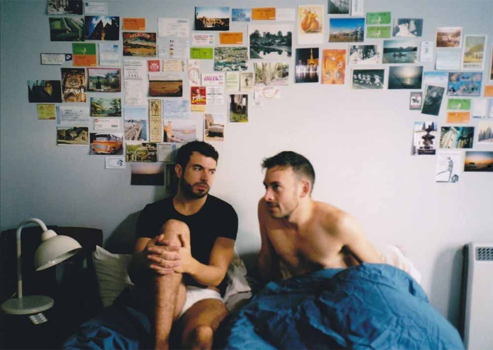 A still from Weekend featuring Russell and Glen, two young white men, sit in bed together, with a vibrant collage of photos on the wall above them. The photo collage was designed by production designer Sarah Finlay.