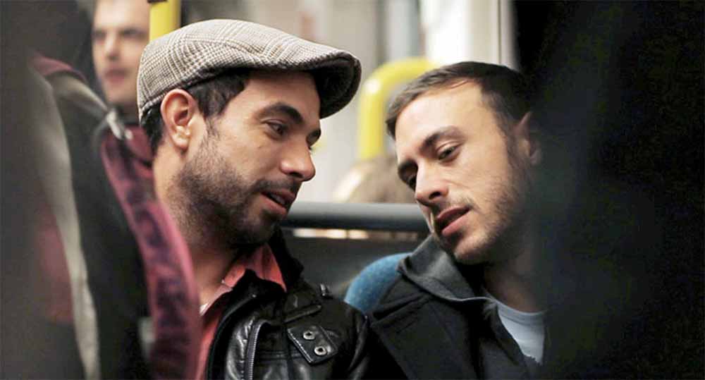A still from Weekend, shot by Ula Pontikos, in which Russell and Glen, two oung white men, sit together on a tram, leaning in toward each the frame crowded by other tram passengers.