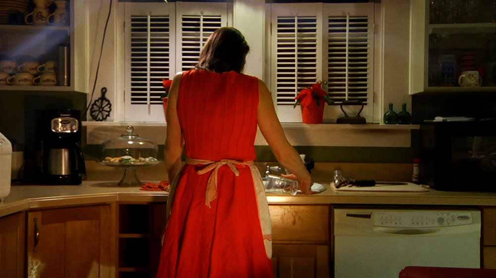A still from Robert Greene's Actress, in which actress Brandy Burre stands with her back to the camera, leaning on her kitchen countertop, wearing a red dress.