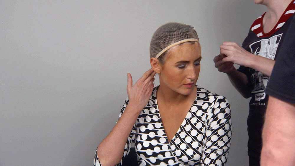 A still from Robert Greene's Kate Plays Christine, in which actress Kate Lyn Sheil sits in a film set with a hair net on her hair, and she holds her fingers to the back of her head, imitating a gun.