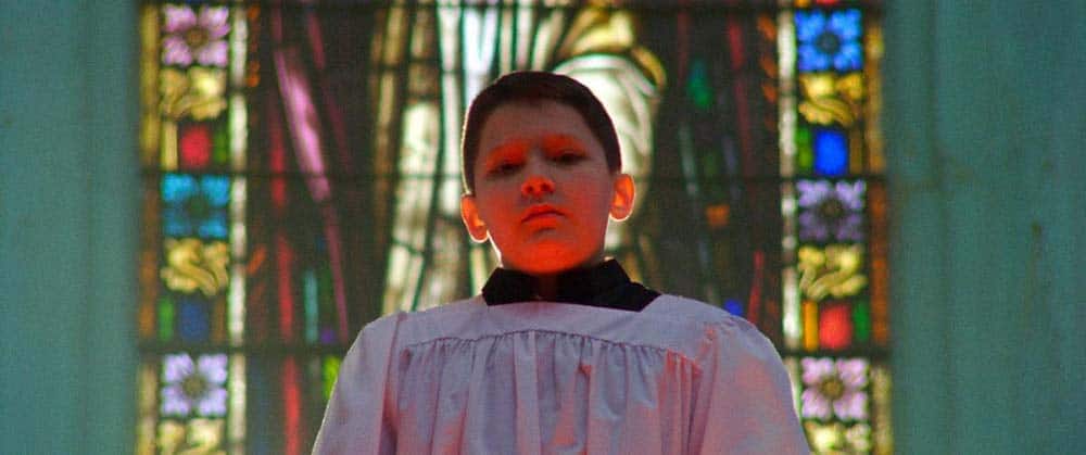 A still from Procession, in which a young boy in church robes stands in front of a stained glass church window, his head haloed with light.