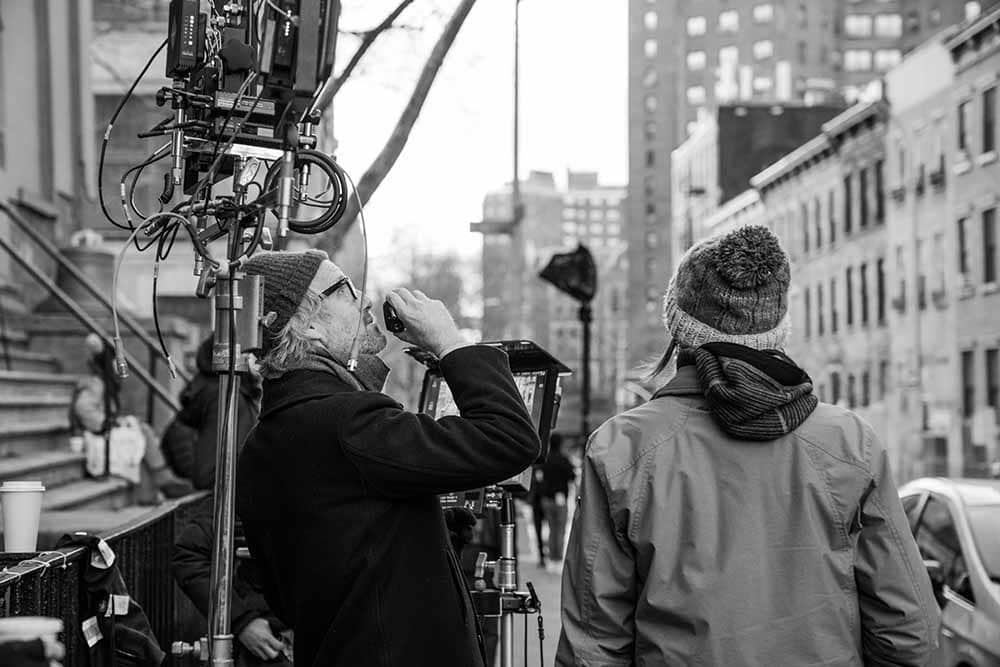 A black and white still from behind the scenes of C'mon C'mon, in which director Mike Mills stands among a load of film equipment, with the New York skyline behind him.