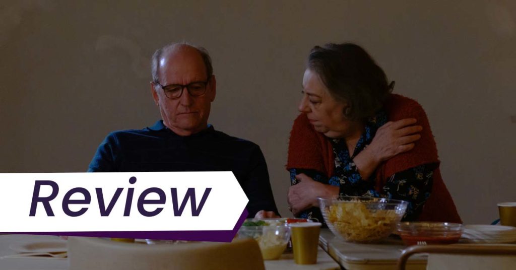 A still from The Humans, in which an aging heterosexual couple, played by Richard Jenkins and Jane Houdyshell, sits at a dinner table, the woman looking pointedly at her husband while her looks down at the food in front of him. The text on the image reads, 'Review'.