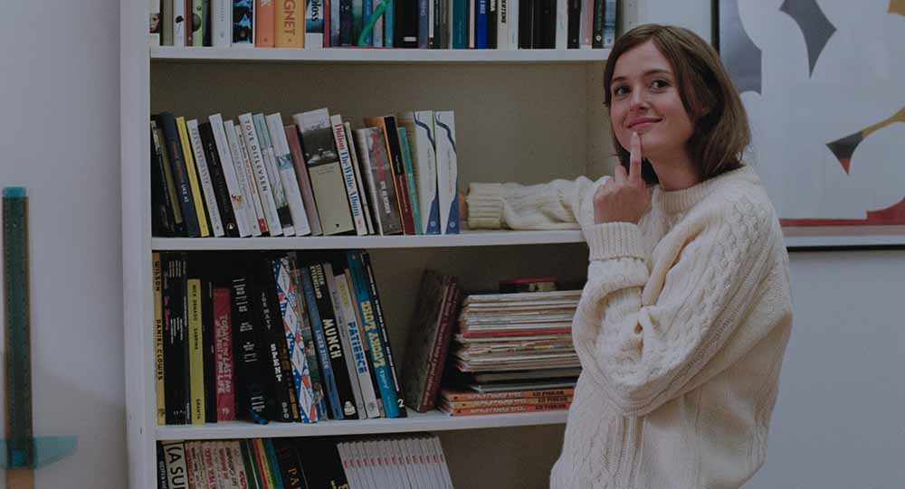 A still from The Worst Person in the World, in which a young woman organises her bookshelf, her fingers up to her mouth in a cheeky, questioning pose.