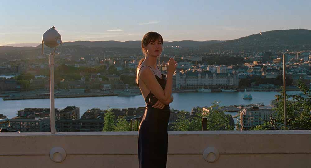 A still from The Worst Person in the World, in which Renate Reinsve as Julie stands on a balcony overlooking Oslo, holding a cigarette in one hand, wearing a slimline dark blue dress.