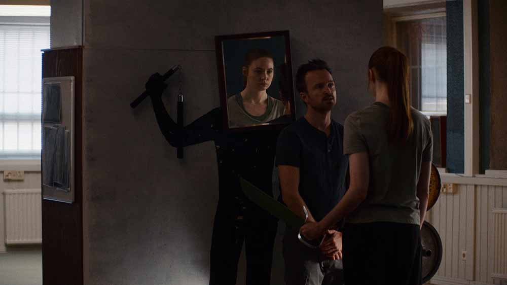 A still from Dual, one of the best films of Sundance 2022, in which a woman stands looking into a mirror that is attached to the head of a dummy. Her expression is downturned, and a man stands in front of her, trying to encourage her.