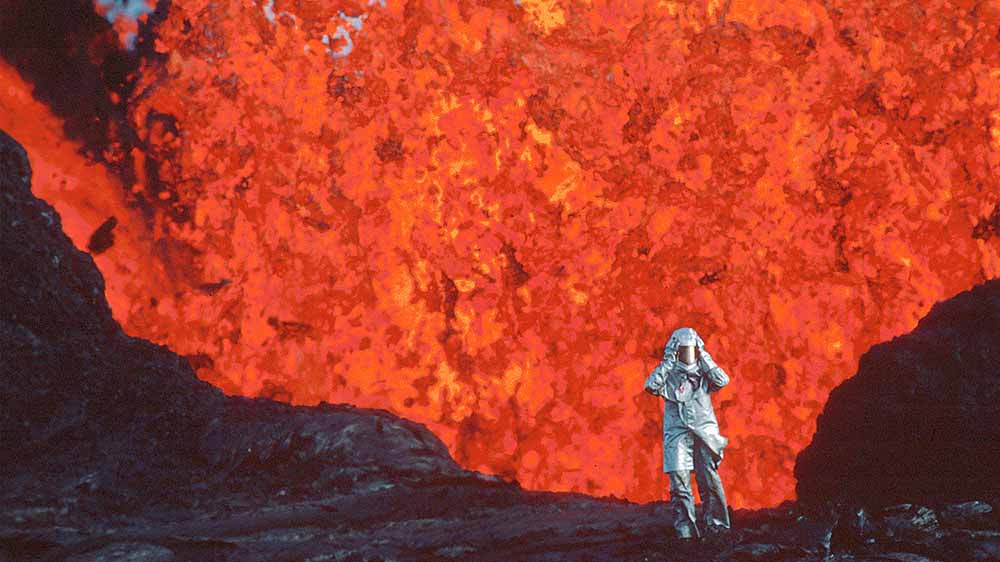 A still from Fire of Love, one of the best films of Sundance 2022, in which a small figure dressed in white protective gear and helmet stands against a vast wall of red lava.