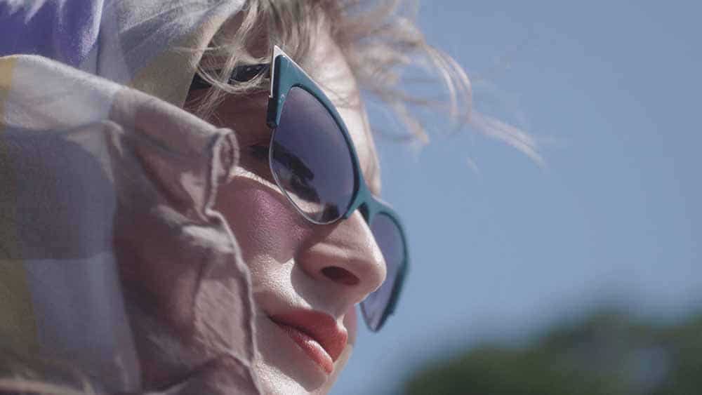 A still from Framing Agnes, which shows a closeup of Agnes's face, wrapped in a headscarf, eyes covered with teal sunglasses, lips red with lipstick, blonde hair whipped up by the wind.