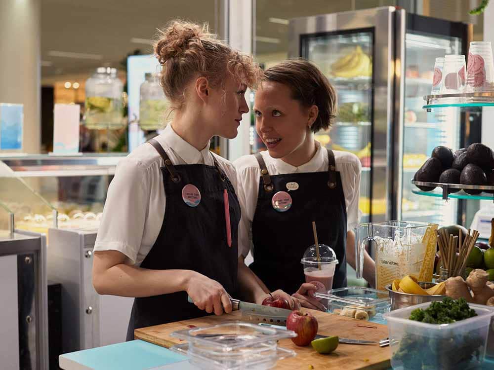 A still in which two teenage girls stand in uniform, which is white shirts and a black apron, for the smoothie stall they work at, laughing hysterically as one of them chops up fruit for a smoothie.
