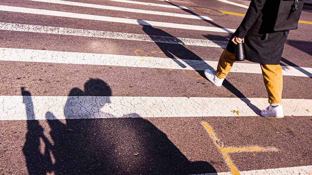 A still from I Didn't See You There, which shows us a slightly slanted view of a crosswalk, featuring the legs of a woman walking across it, and the shadow of a man in a wheelchair.