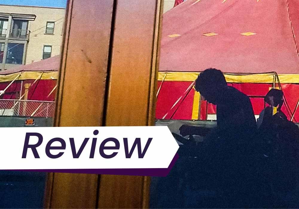 A still from I Didn't See You There, in which a man in a wheelchair is seen silhouetted as a reflection in a glass panel, his silhouette eclipsed by the looming figure of a red and yellow circus tent. The text on the image reads, 'Review'.