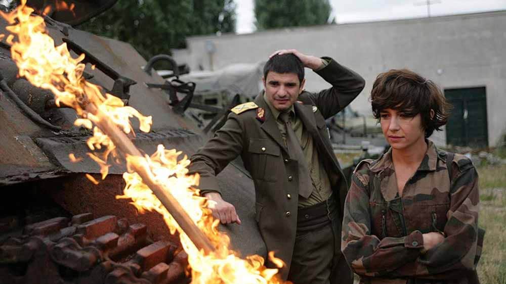 A man and a young woman dressed in military uniform stand by a pile of burning wood, in this still from I Do Not Care If We Go Down in History as Barbarians.