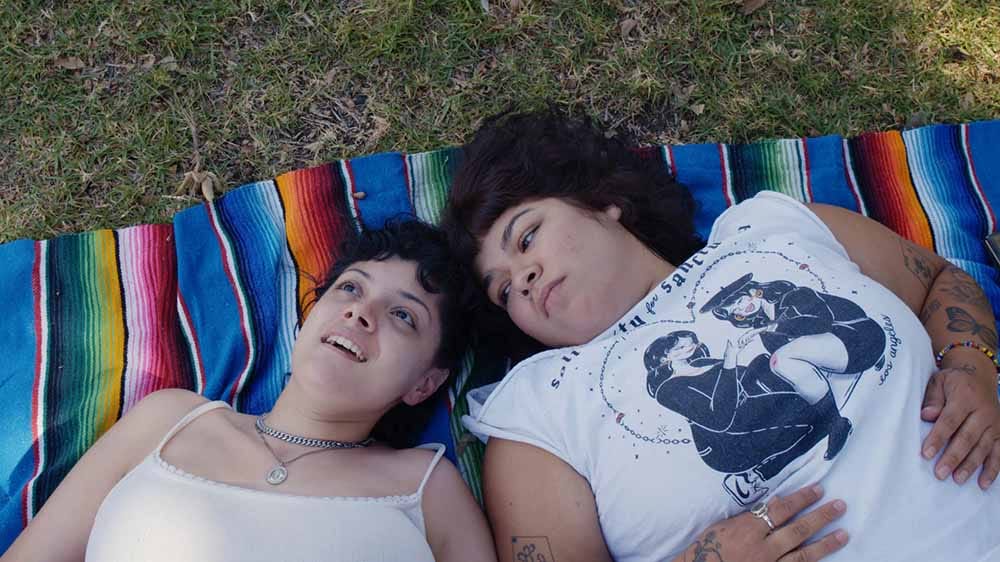 A still from Mija, in which two young Mexican-American women, Jacks and Doris, lie down on a multicoloured picnic blanket. Jacks stares at the sky while Doris stares as Jacks.