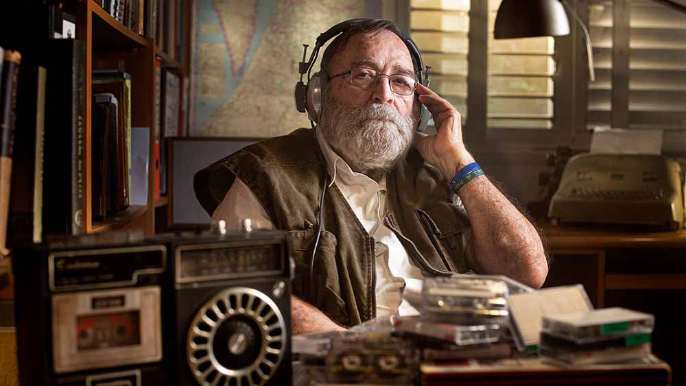 A still from Tantura, one of the best films of Sundance 2022, in which an older man listens to a tape on a pair of headphones, one hand to his ear, while he stares gravely into the camera.