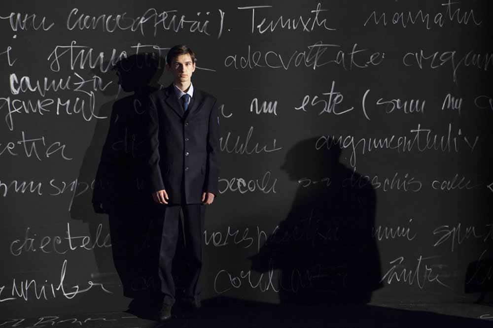 A still from Uppercase Print, directed by Radu Jude, in which a slim young man in a suit stands in front of a black chalkboard scrawled with words.