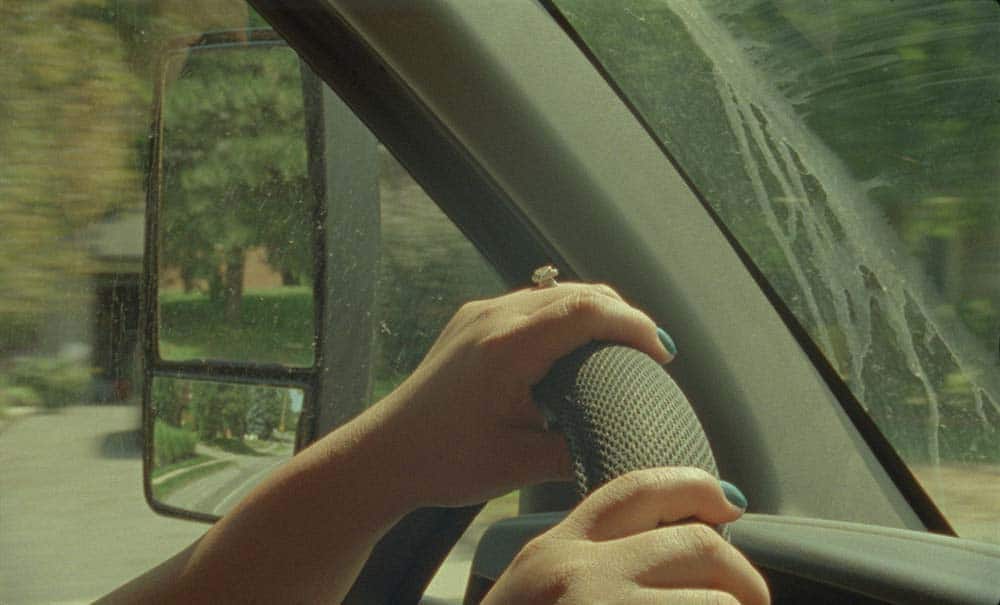 A still from My Two Voices, picturing a pair of hands resting on a car wheel, with soft sunlight highlight the backs of her hands.