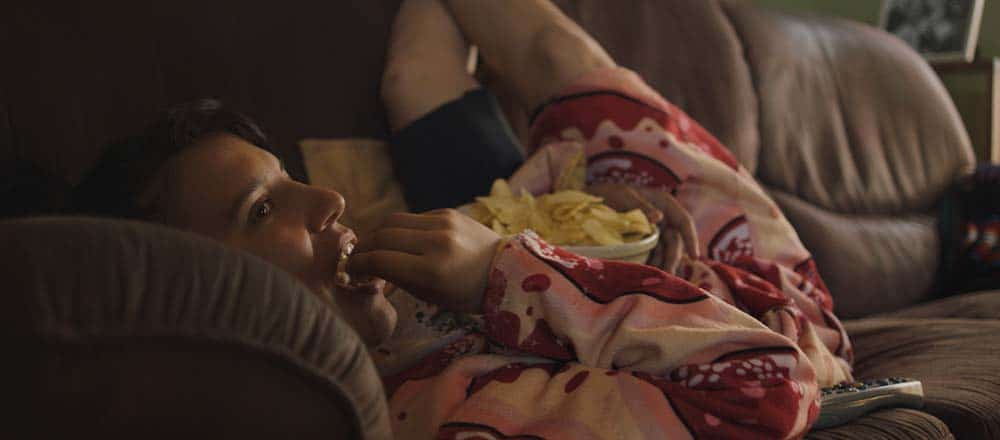 A still from Run Woman Run, in which main character Beck lies on the sofa eating from a bowl of chips.