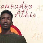 A still of actor Mamoudou Athie, in front of a yellow, orange, and red background, and text that displays his name.